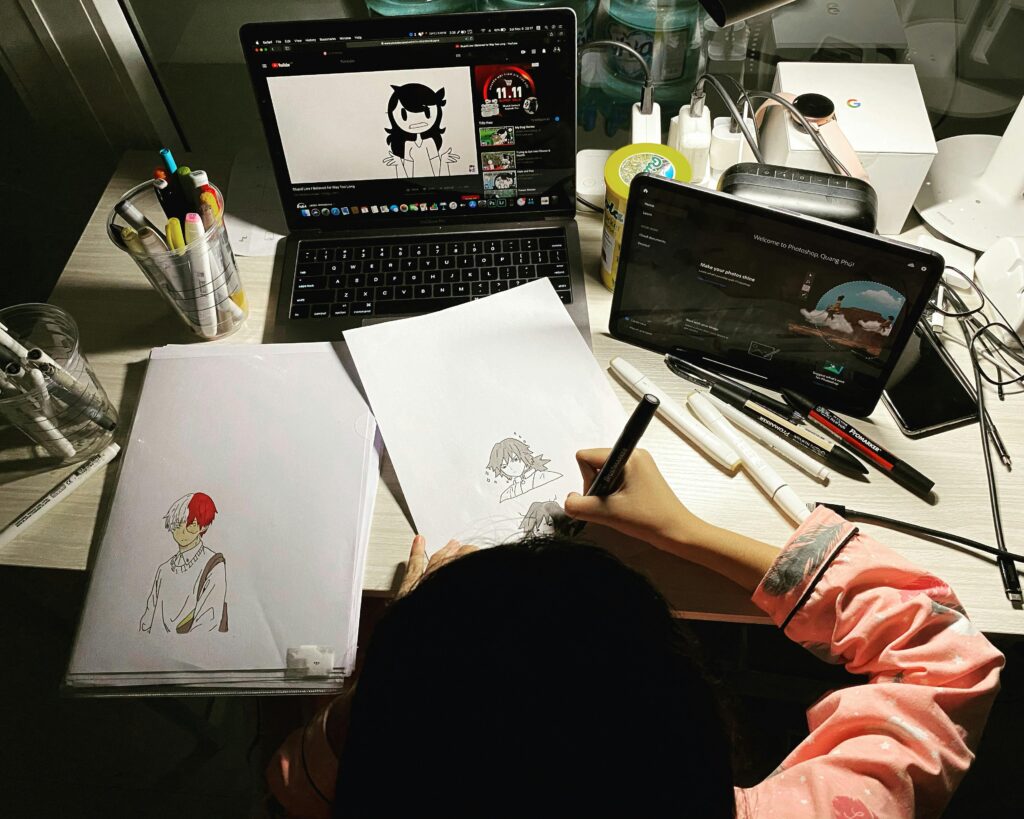 Artist sketching anime characters while watching a tutorial on a laptop, surrounded by artistic tools.